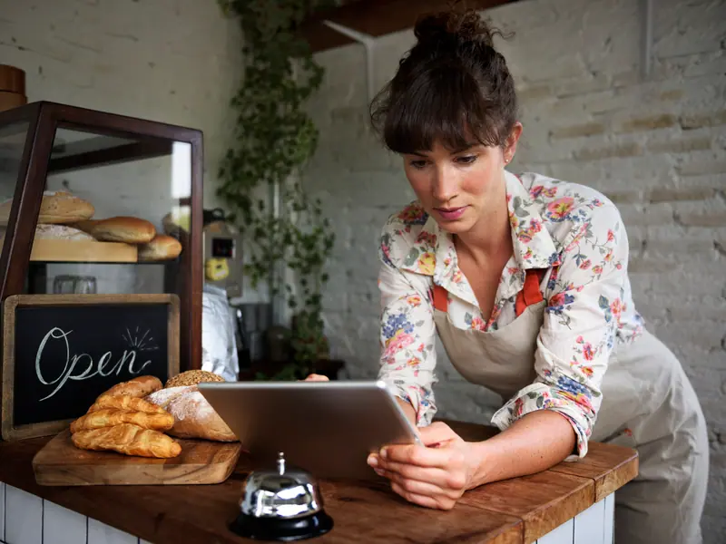 Seorang pembuat roti berkemeja motif bunga dan bercelemek membaca tablet di meja dengan roti segar dan papan bertuliskan Open.