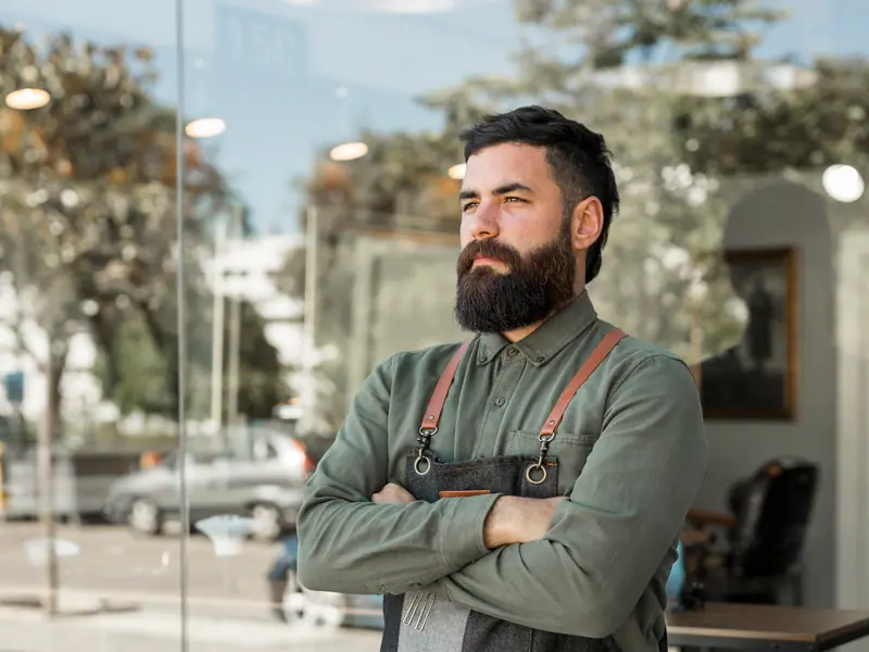 Seorang barber berjanggut dengan celemek denim berdiri bersedekap di depan jendela toko.