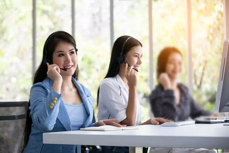 Tiga agen dukungan memakai headset menerima panggilan di meja bersama dekat jendela yang terang.